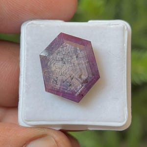 May include: A faceted, hexagonal gemstone with a deep purple hue, set in a white plastic case. The gem's interior features intricate, light-colored patterns. The background is blurred, suggesting an outdoor setting.
