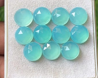May include: A collection of ten round, faceted, light blue gemstones. Each gem has a domed top with multiple facets that catch the light. The gems are arranged in a white tray, with a hand holding the tray.