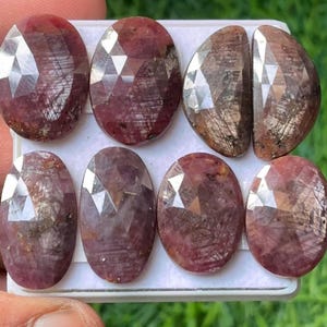 Op de afbeelding: Acht facetgeslepen ovale edelstenen in rood en zilver. De edelstenen zijn gerangschikt in twee rijen van vier op een wit oppervlak.