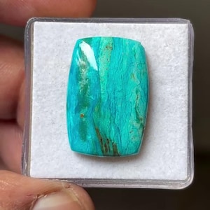 May include: A rectangular cabochon gemstone featuring a vibrant turquoise and teal color palette with natural dark brown veining and inclusions. The stone has a polished, reflective surface and is displayed inside a square clear plastic case.