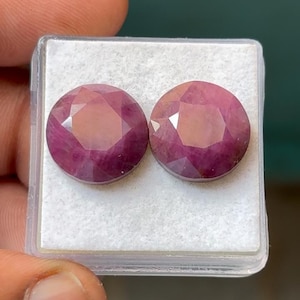 May include: Two round, faceted gemstones with a deep pink hue are displayed in a clear plastic case. The gems have multiple facets that catch the light, creating a sparkling effect. The case sits on a white surface.
