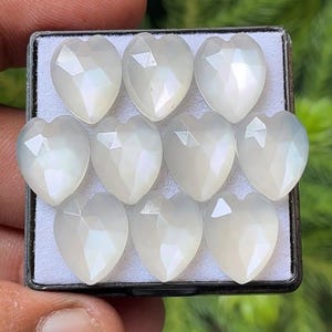 Może przedstawiać: Kolekcja dziesięciu fasetowanych kamieni szlachetnych w kształcie serca. Kamienie mają perłowo-biały kolor i są ułożone w czarnym prostokątnym pojemniku. Fasetki tworzą efekt połysku.