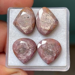 May include: Four faceted, pink-brown gemstones in a clear plastic case. Two are teardrop-shaped, and two are diamond-shaped. The gems have internal inclusions and a polished, reflective surface. The gems are likely for jewelry making.