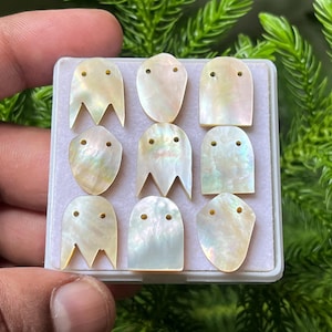May include: A collection of nine iridescent mother-of-pearl shell components arranged in a three-by-three grid on a white display card. Each piece features a smooth, pearlescent surface with two small drilled holes for attachment or threading.