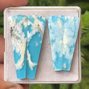 May include: Two polished gemstone slabs with a striking blue and white cloud-like pattern. The stones are set in a white tray, showcasing their unique natural designs. The gemstones have a polished, glossy finish.