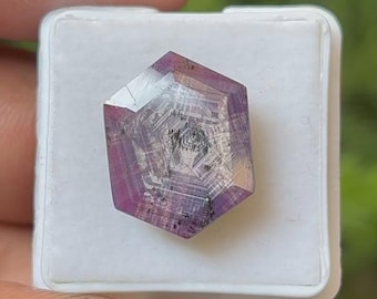 Op de afbeelding: Een gefacetteerde, zeshoekige edelsteen met een paarse tint, geplaatst in een witte plastic container. De edelsteen heeft interne patronen en insluitsels, met heldere en ondoorzichtige gebieden. De achtergrond is vaag groen.
