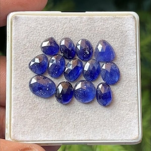 May include: A collection of faceted, deep blue gemstones displayed in a white rectangular container. The gemstones have various shapes, including oval and triangular, and reflect light with multiple facets. The container is held by a hand.