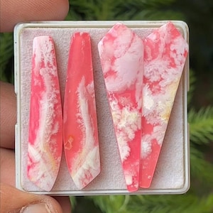 May include: Five polished gemstones in a white tray. The stones are a vibrant pink with white and yellow cloud-like patterns. The shapes vary from rectangular to triangular, showcasing natural stone variations.