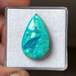 May include: A teardrop-shaped turquoise gemstone cabochon featuring a vibrant mosaic of bright teal and cyan fragments with a polished, glossy surface. The stone displays varied inclusions and a distinct dark blue leaf-like pattern embedded within.