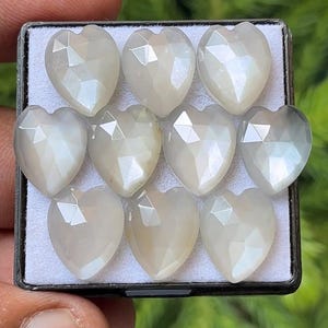 Może przedstawiać: Kolekcja dziesięciu kamieni szlachetnych w kształcie serca, każdy z fasetowaną powierzchnią, prezentowanych w czarnej tacy. Kamienie mają perłową, opalizującą jakość, odbijającą światło. Ogólny kolor to miękki, złamany biały.