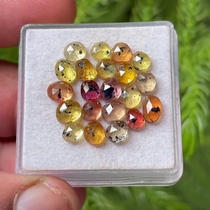 Puede incluir: Una colección de pequeñas piedras preciosas facetadas y multicolores dispuestas en una caja de presentación cuadrada. Las piedras presentan varios tonos de amarillo, naranja y rosa, con inclusiones oscuras visibles y una superficie pulida.