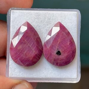 May include: Two faceted, teardrop-shaped rubies in a clear plastic case. The rubies are a deep pinkish-red color with internal striations and a high luster. One ruby has a small, dark inclusion. The gems are displayed against a white background.