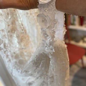 May include: Close-up of a white wedding dress detail. The dress features delicate lace, small pearls, and white fabric flowers. The dress is held up, showcasing the intricate design and texture of the fabric.