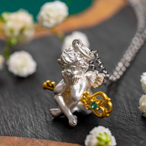Op de afbeelding: Zilveren cherubijn hanger ketting met een gouden sleutel en een blauwe edelsteen. De cherubijn houdt de sleutel vast en heeft zijn hoofd op zijn hand rusten.