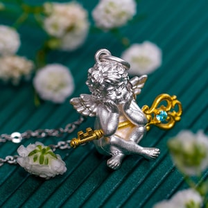 Op de afbeelding: Een zilveren cherub-hanger met een gouden sleutel en een blauwe edelsteen. De hanger is aan een zilveren ketting en is omringd door witte bloemen.