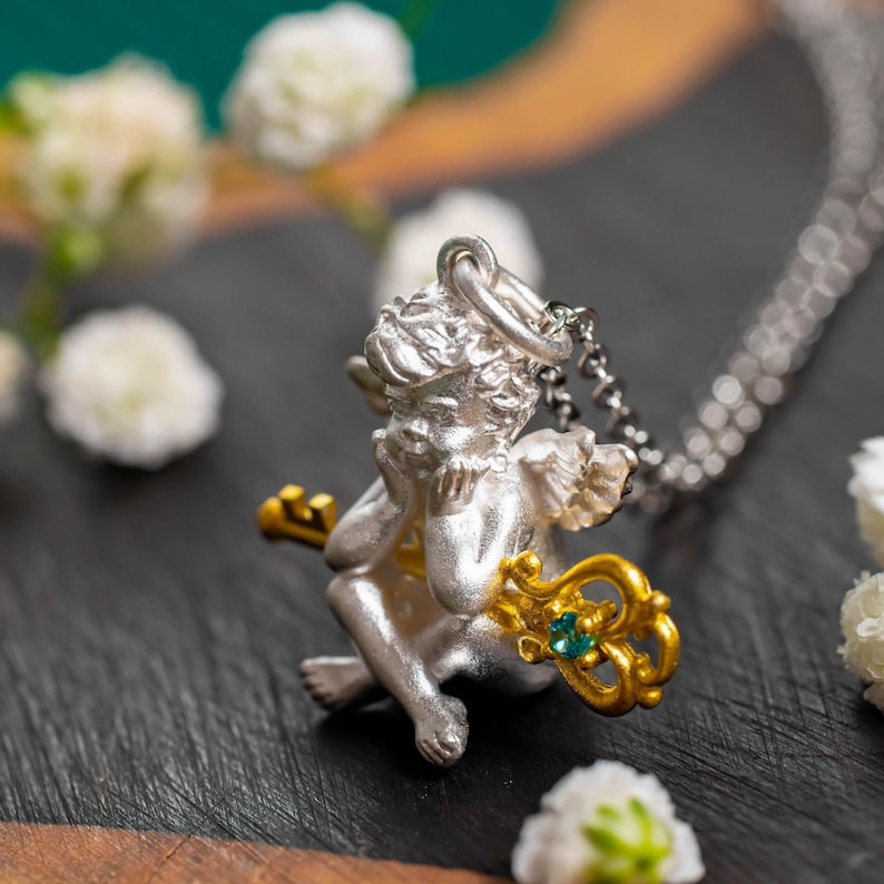 Op de afbeelding: Zilveren cherubijn hanger ketting met een gouden sleutel en een blauwe edelsteen. De cherubijn houdt de sleutel vast en heeft zijn hoofd op zijn hand rusten.