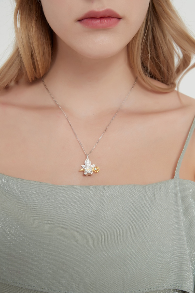 Op de afbeelding: Een zilveren ketting met een delicate hanger met een gestileerde engel die een sleutel vasthoudt. De engel is gemaakt van zilver en goud, en de sleutel is van goud. De hanger heeft een kleine blauwe edelsteen.