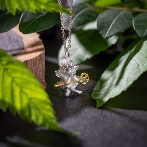 Op de afbeelding: Zilveren en gouden ketting met een hanger in de vorm van een cherubijn met een blauwe edelsteen sleutel. De ketting wordt getoond op een donkere achtergrond met groene bladeren.