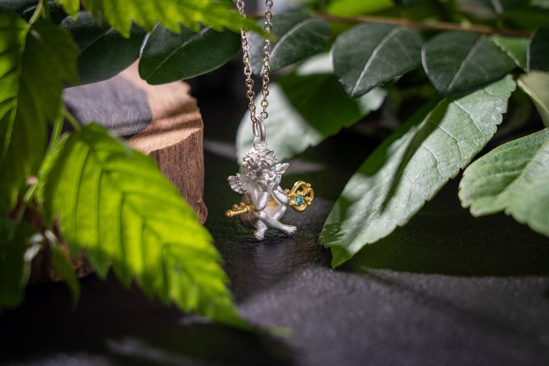 Op de afbeelding: Zilveren en gouden ketting met een hanger in de vorm van een cherubijn met een blauwe edelsteen sleutel. De ketting wordt getoond op een donkere achtergrond met groene bladeren.