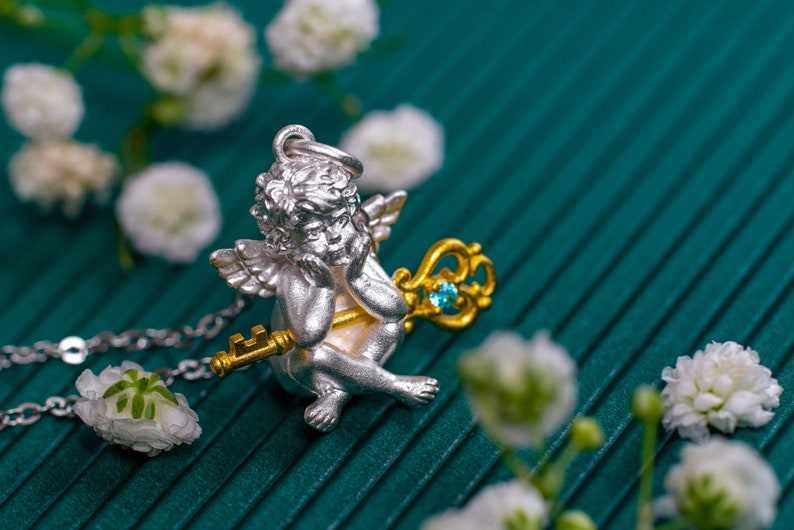 Op de afbeelding: Een zilveren cherub-hanger met een gouden sleutel en een blauwe edelsteen. De hanger is aan een zilveren ketting en is omringd door witte bloemen.