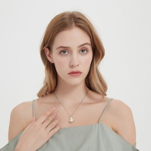 Op de afbeelding: Een vrouw draagt een lichtgroen top met off-shoulder, een zilveren kettingketting en een klein hanger. De hanger is een zilveren bloem met een parel in het midden.