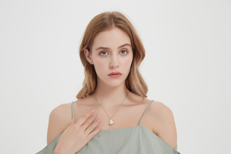 Op de afbeelding: Een vrouw draagt een lichtgroen top met off-shoulder, een zilveren kettingketting en een klein hanger. De hanger is een zilveren bloem met een parel in het midden.