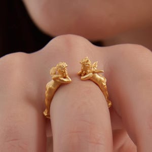 May include: A gold ring with two cherub figures. The cherubs have detailed wings and are posed as if resting. The ring is worn on a finger, highlighting the design's intricate details and craftsmanship.