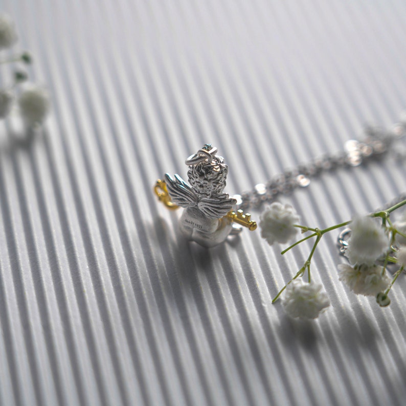 Op de afbeelding: Zilveren ketting met een klein engelbedeltje dat een sleutel vasthoudt. De engel draagt een halo en heeft vleugels. De ketting ligt op een witte, textuurrijke achtergrond met kleine witte bloemen.