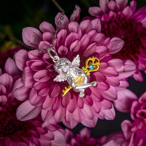 Op de afbeelding: Een zilveren cherub-hanger met een gouden sleutel en een blauwe edelsteen, hangend aan een zilveren ketting. De hanger rust op een tros roze bloemen.