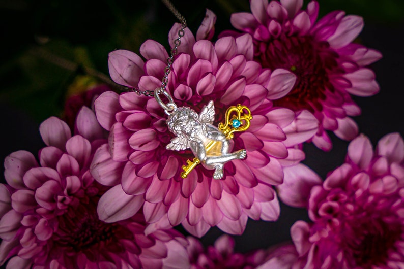 Op de afbeelding: Een zilveren cherub-hanger met een gouden sleutel en een blauwe edelsteen, hangend aan een zilveren ketting. De hanger rust op een tros roze bloemen.