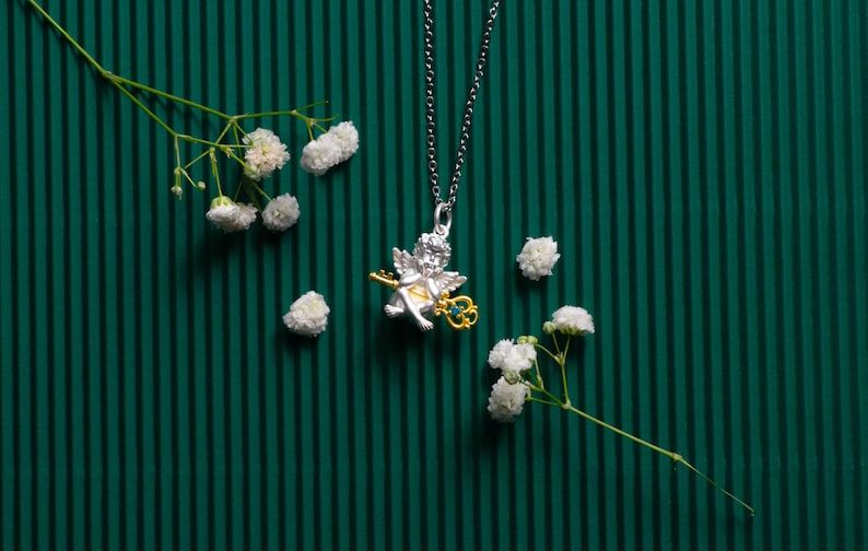 Op de afbeelding: Zilveren ketting met een gouden cherubijn hanger die een sleutel vasthoudt. De ketting ligt op een groene gestreepte achtergrond met witte bloemen.