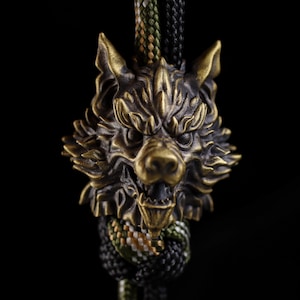 Puede incluir: Una cuenta de metal en tono bronce con forma de cabeza de lobo, con detalles intrincados de pelaje y expresión de boca abierta. La cuenta está ensartada en un cordón grueso de paracord trenzado con un patrón de camuflaje verde, negro y beige.