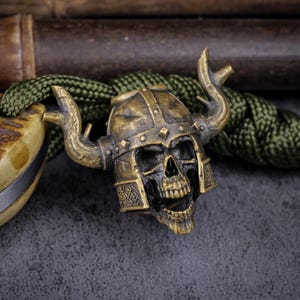 May include: A bronze-colored skull bead with a horned helmet, detailed with rivets and facial features. The bead is attached to a green cord, with a portion of a knife handle visible on the left. The background is a dark, textured surface.
