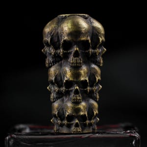May include: A bronze-colored, stacked skull sculpture. The sculpture is composed of multiple skulls arranged in a vertical column. The skulls have detailed features and are set against a dark background. The sculpture is a decorative item.