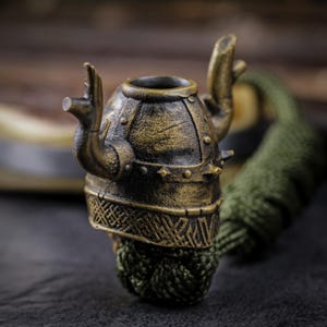 May include: A detailed, antique-style pipe with a bronze finish. The pipe is shaped like a Viking helmet, featuring intricate details and a green, braided stem. The design includes raised hand-like elements and decorative patterns.