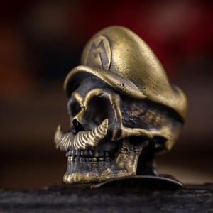 May include: A metallic brass-toned ring featuring a sculpted human skull wearing a cap with a prominent letter M on the front. The skull has a detailed moustache and a weathered, dark finish that highlights the textured contours of the metal design.
