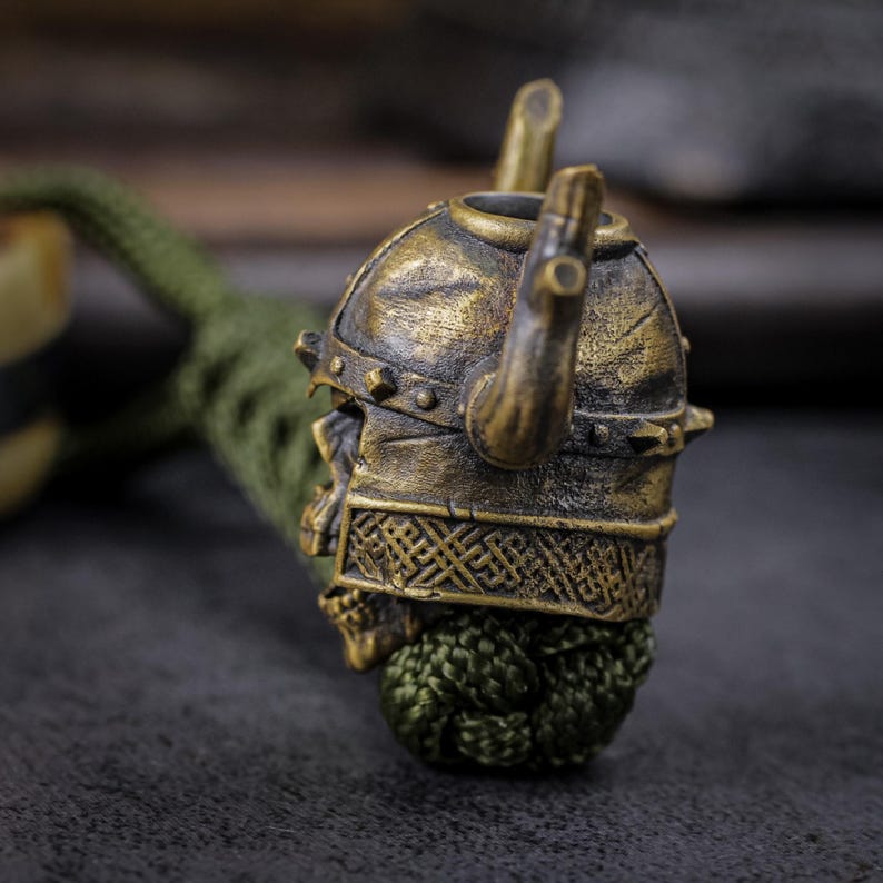 May include: A bronze-colored Viking helmet pendant with horns and intricate detailing. The helmet is attached to a green cord. The pendant has a skull-like appearance. The background is a dark, textured surface.