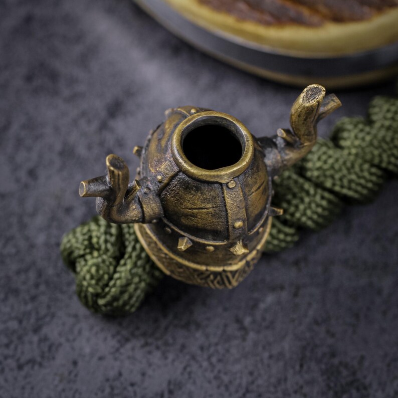 May include: A bronze-colored, decorative bottle stopper with a unique, helmet-like design. The stopper features a cylindrical body with a wide opening and is adorned with rivets and spikes. It rests on a green, braided cord against a dark gray background.