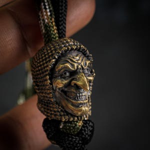 May include: A bronze-colored pendant featuring a detailed, grinning witch's face with a pointed nose and hooded cowl. The pendant is attached to a black and green braided cord, creating a unique accessory.