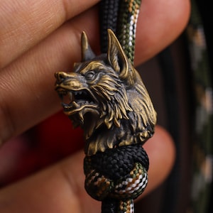 Puede incluir: Una cuenta con forma de cabeza de lobo en bronce metálico con textura de pelaje detallada y una expresión de boca abierta con dientes visibles. La cuenta está ensartada en un cordón de paracord grueso y trenzado con un patrón de camuflaje.