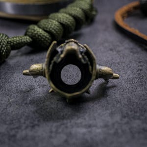 May include: A detailed close-up of a bronze-colored metal object with a central hole. The object has an intricate design with small protrusions and is set against a dark gray surface. The object appears to be a decorative element.