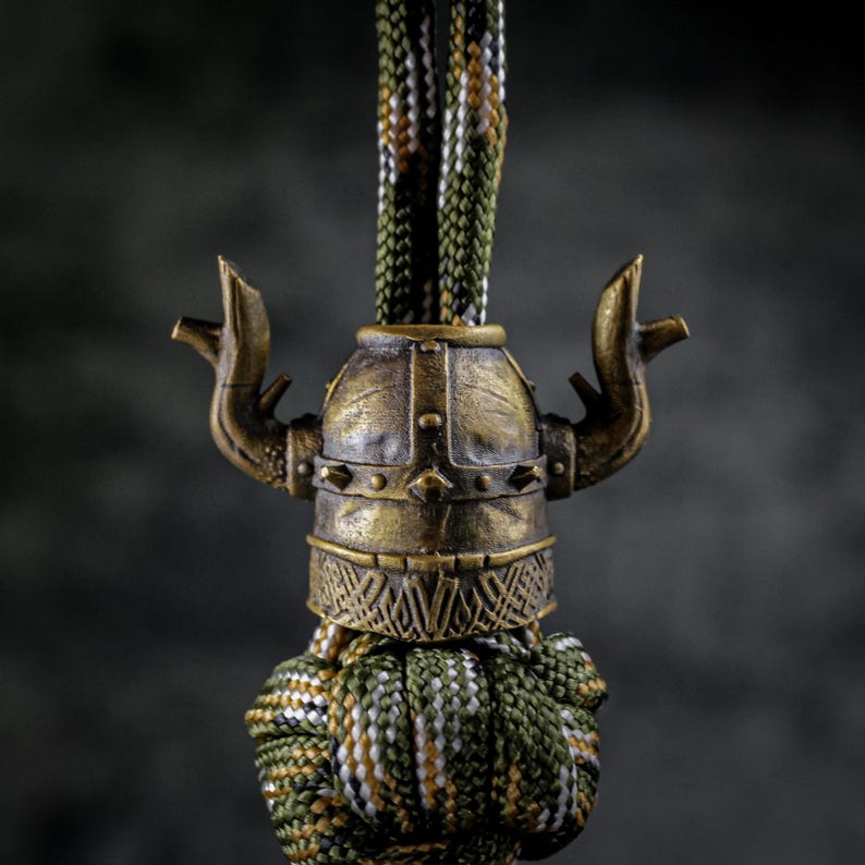 May include: A bronze-colored Viking helmet charm with intricate detailing, including horns and rivets, is attached to a braided cord in shades of green, brown, and white. The charm has an aged, antique appearance.