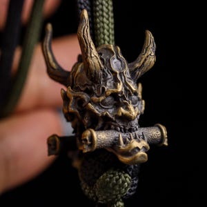 May include: A bronze-toned metal pendant featuring a detailed horned demon mask with a textured surface. The demon is holding a small cylindrical scroll in its mouth. The pendant is attached to a thick, braided olive green and black cord.