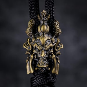 May include: A bronze-coloured pendant with a detailed demon face design, suspended from a black braided cord. The pendant features horns, ornate facial details, and a crown-like element. The cord is made of a black, braided material.