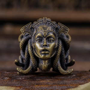 May include: A bronze-colored Medusa head sculpture with intricate details. The sculpture features a woman's face with snakes for hair, a crown, and a pendant. The piece has a weathered appearance, suggesting an antique or vintage style.