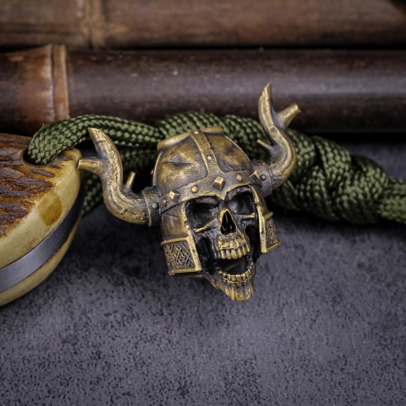 May include: A bronze-colored skull bead with a horned helmet, detailed with rivets and facial features. The bead is attached to a green cord, with a portion of a knife handle visible on the left. The background is a dark, textured surface.