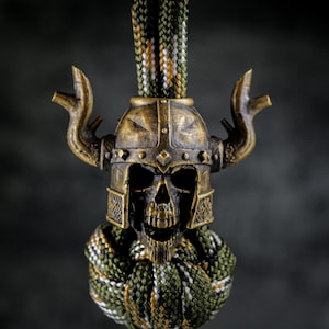 May include: A bronze-colored skull wearing a Viking helmet with horns. The skull is detailed with visible teeth and eye sockets. The helmet has intricate designs. A green, white, and brown braided cord is attached to the skull.