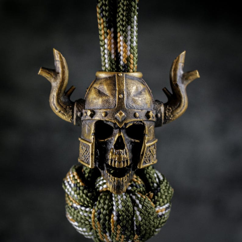 May include: A bronze-colored skull wearing a Viking helmet with horns. The skull is detailed with visible teeth and eye sockets. The helmet has intricate designs. A green, white, and brown braided cord is attached to the skull.