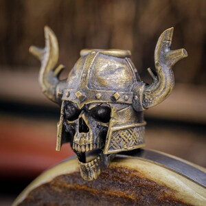 May include: A detailed, antique-style skull figurine wearing a horned Viking helmet. The skull is dark with a bronze-colored helmet, featuring intricate details and a weathered appearance. The horns curve upwards, adding to the Viking aesthetic.