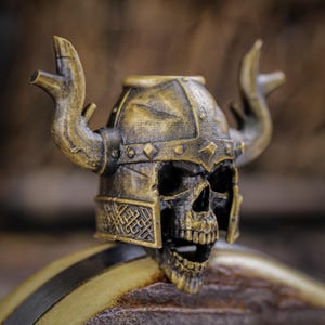May include: A detailed, antique-style skull figurine wearing a Viking helmet with horns. The skull is black with gold accents, and the helmet is a weathered gold color. The figurine has intricate details and a textured finish.